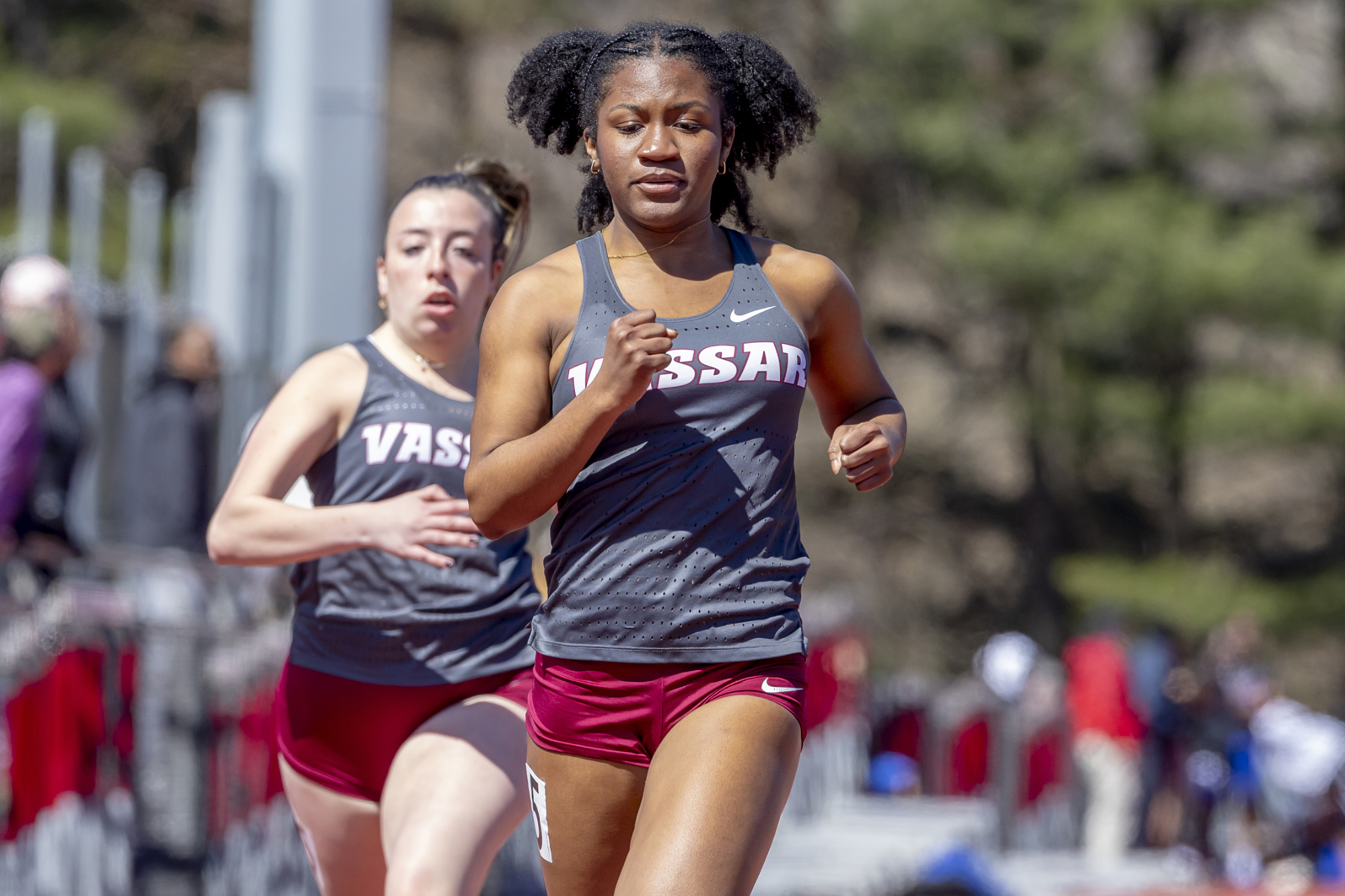 A person with a gray tank top and burgundy shorts runs on a track with a teammate running close behind. The tank top has "Vassar" on it in white letters.