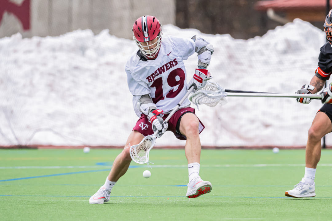 A person wearing a red lacrosse helmet and a white team jersey with "Brewers 19" on it holds a lacrosse racket, going for a ball.