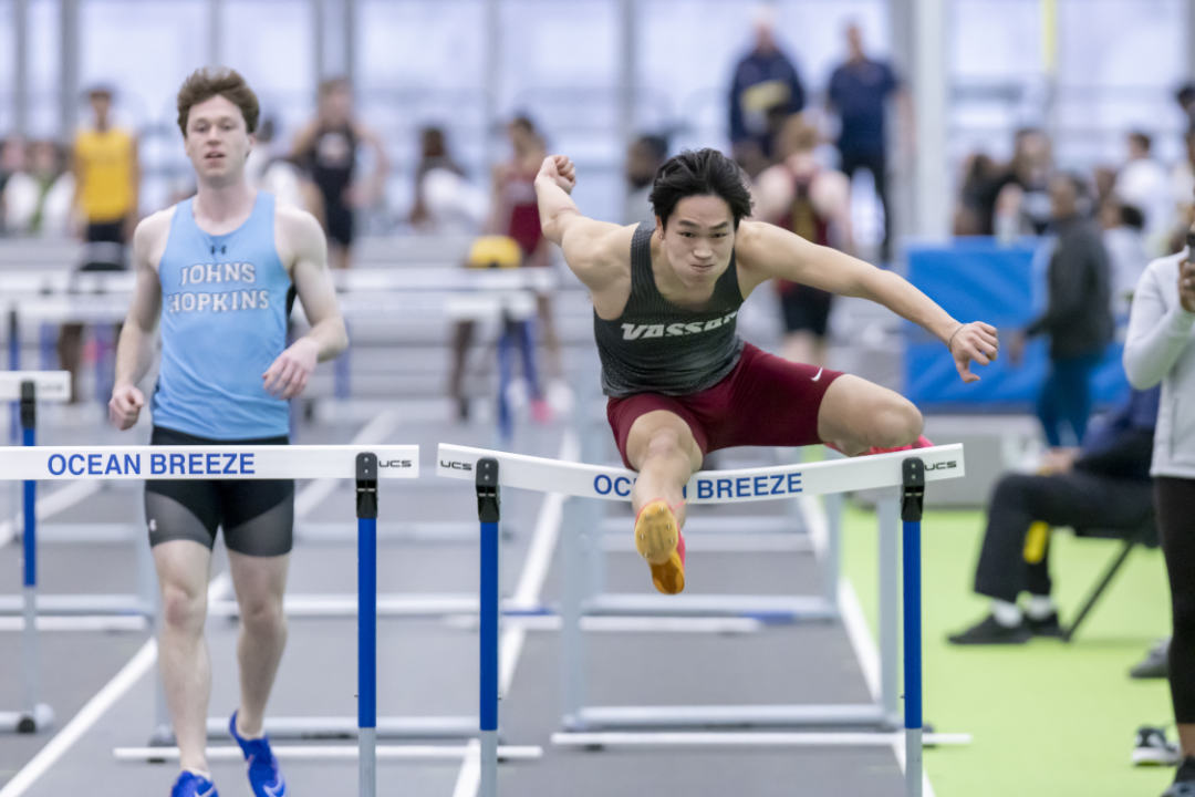 A person wearing red shorts and a black tank top with "Vassar" on it leaps over a barrier with "Ocean Breeze" printed on it.