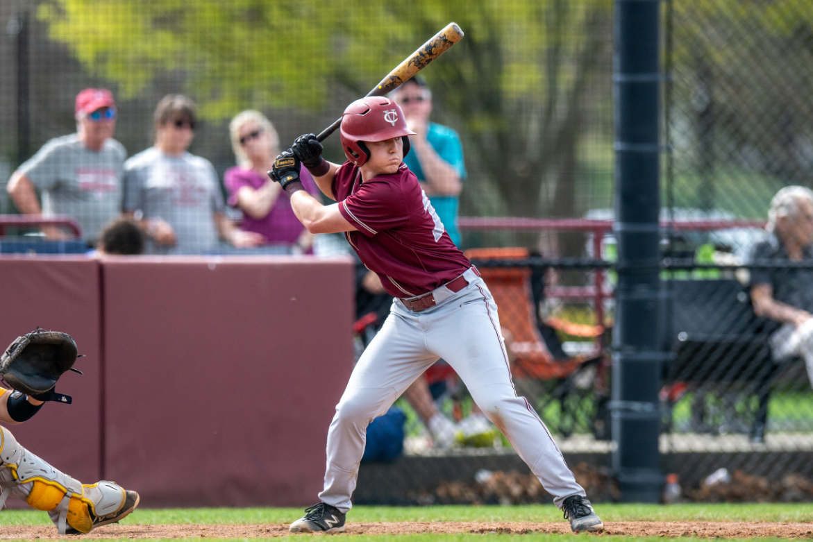 A person wearing light gray pants, a maroon jersey, and a maroon batting helmet winds up to swing at a baseball. The ball is not visible.