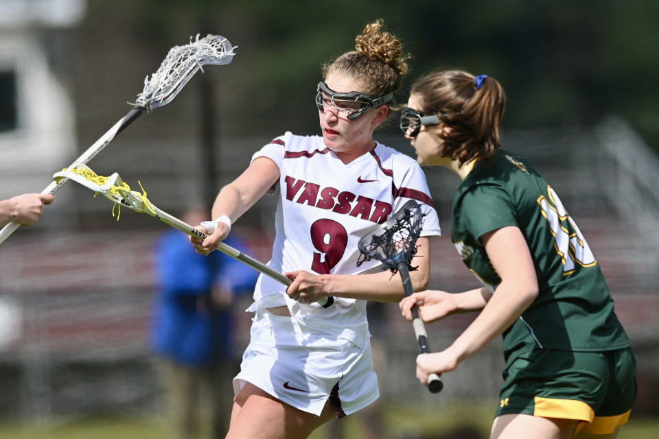 Two lacrosse players from opposing teams. The Vassar player is on the left, and is wearing a white shirt with "Vassar 9" on it. The other player has a dark green shirt and dark green shorts.