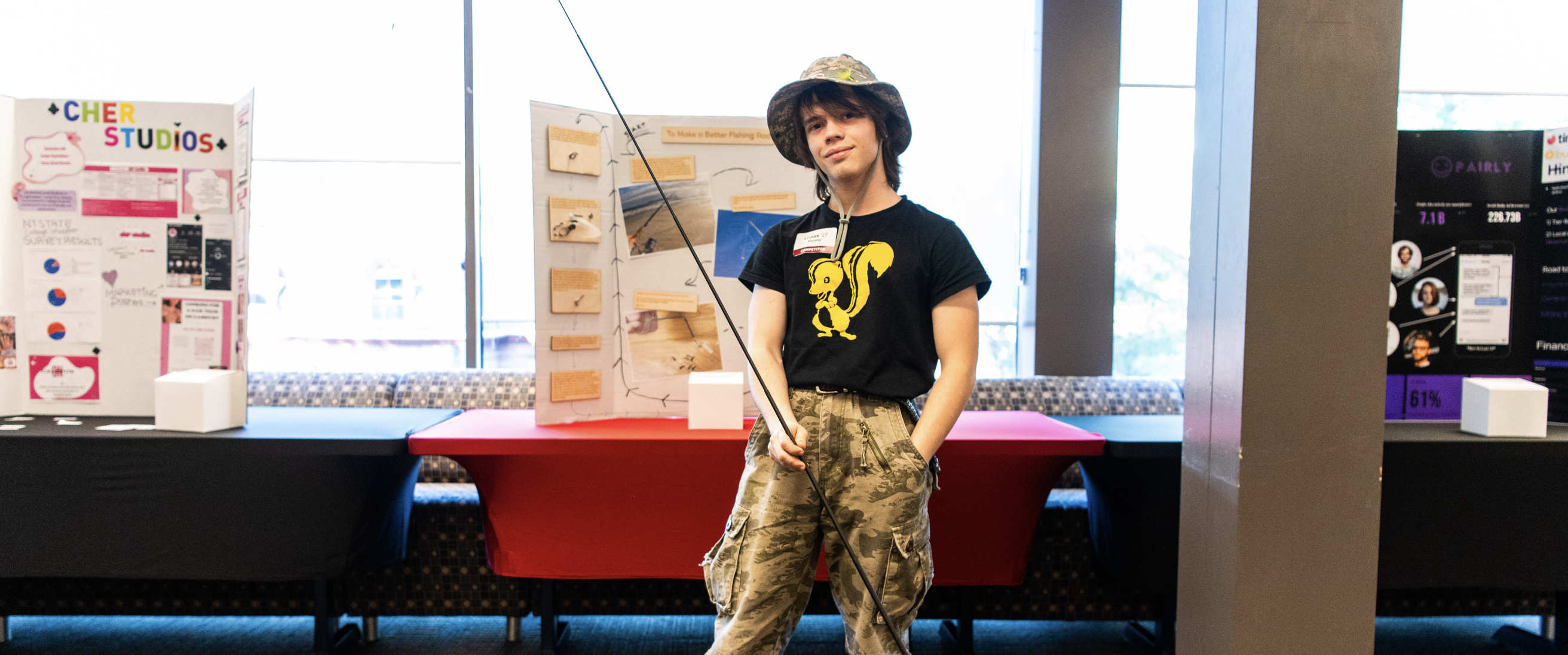 A person with long brown hair stands in front of a wall of large windows. The person is wearing a brimmed hat, camouflage pants, and a black T-shirt with a yellow cartoon drawing of a skunk on it. The person is holding a long fishing pole.