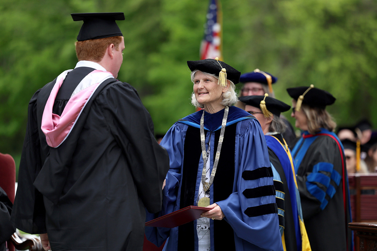 Wearing blue academic regalia, black cap, and a medallion, the College president shakes hands with a graduate during an outdoor ceremony. Faculty members are in the background.