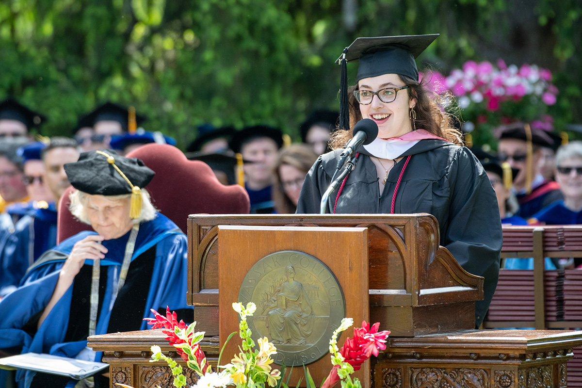 A young person in academic regalia speaks at a podium during an outdoor ceremony.