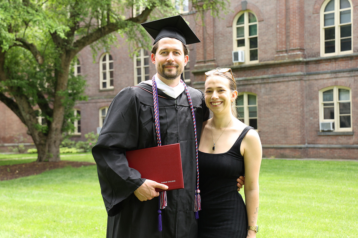 A graduate in a cap and gown holding a diploma stands next to a smiling person on a campus lawn with a brick building in the background.