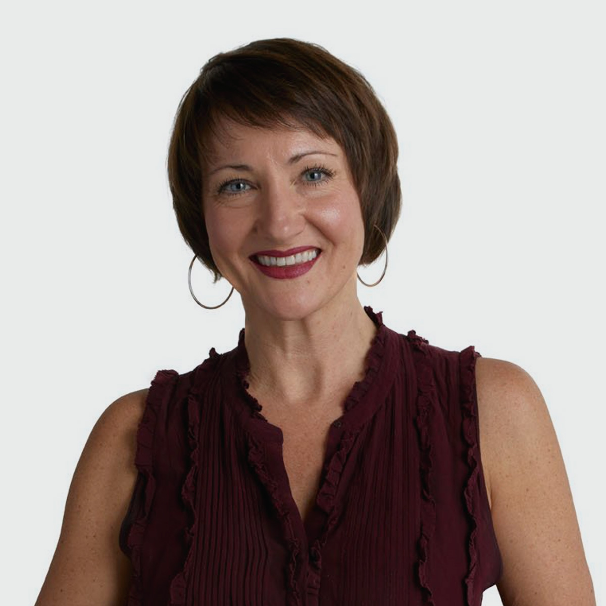 A person with short brown hair is smiling at the camera. They are wearing large hoop earrings and a sleeveless burgundy blouse with a ruffled neckline. 