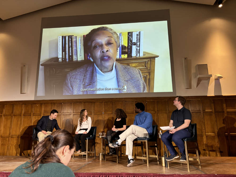 A group of students sit on a stage, above which is projected an image. The image shows a person speaking, and seems to be part of a video.