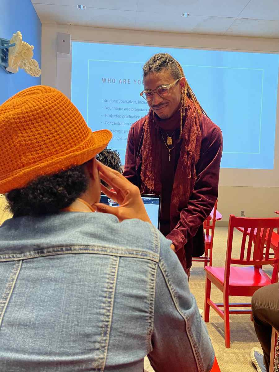 A person with glasses, a short mustache, and long dreadlocks stands in a classroom smiling and listening to someone else speak.
