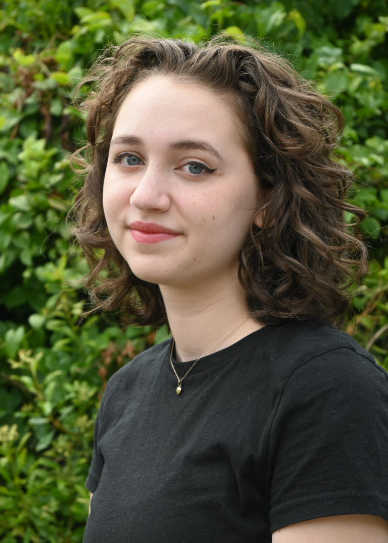 A person standing outside in front of green bushes. The person has long, curly, brown hair and a black shirt.