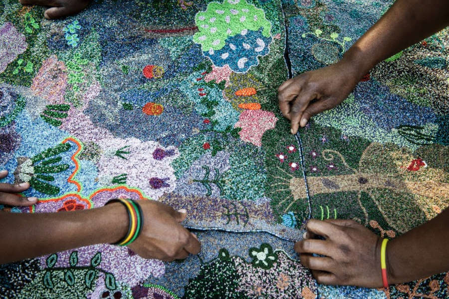 A large, colorful beaded tapestry. Two people are working on it. Only their hands are visible.