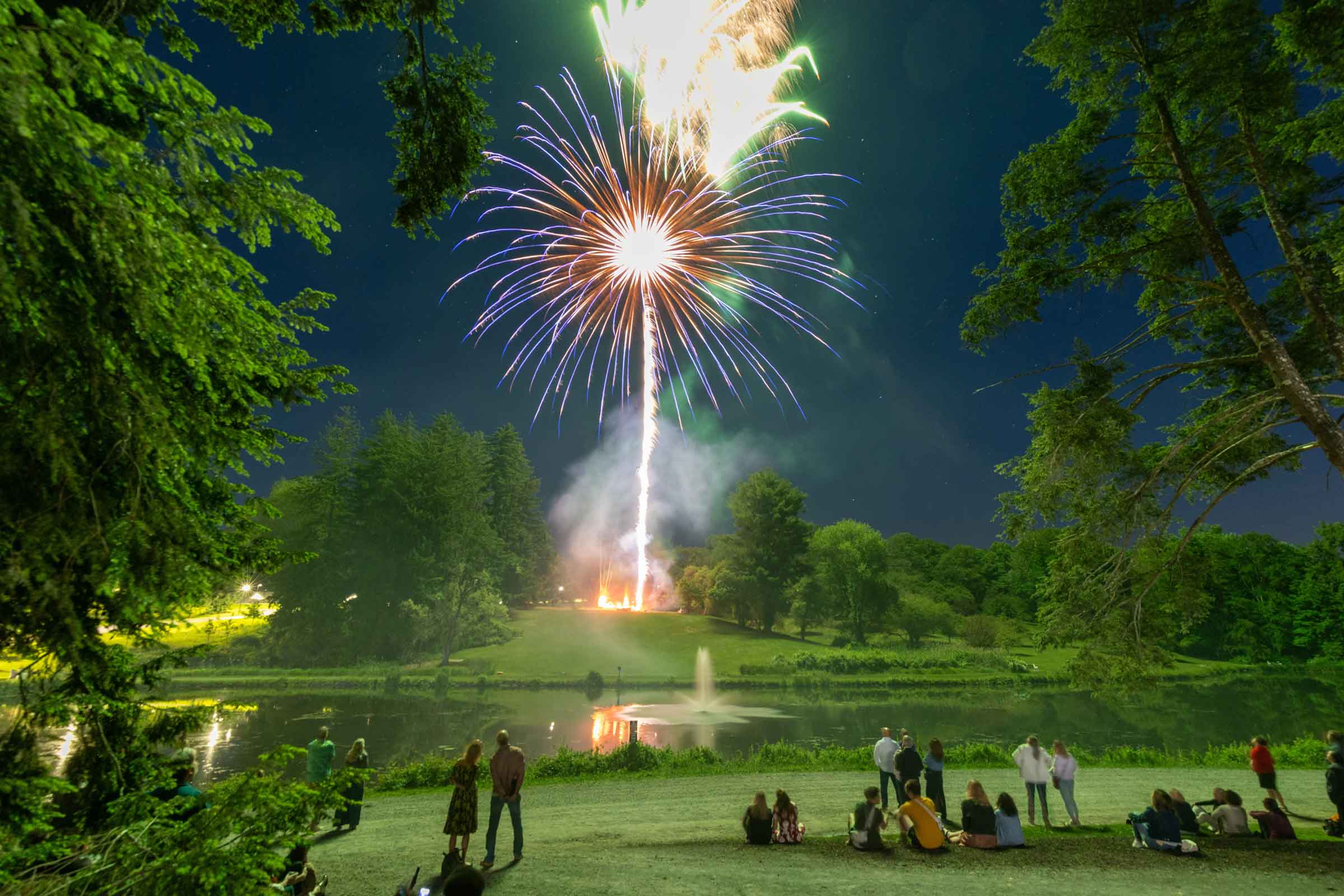 Firework bursting over a lake with onlookers gathered on the shore.