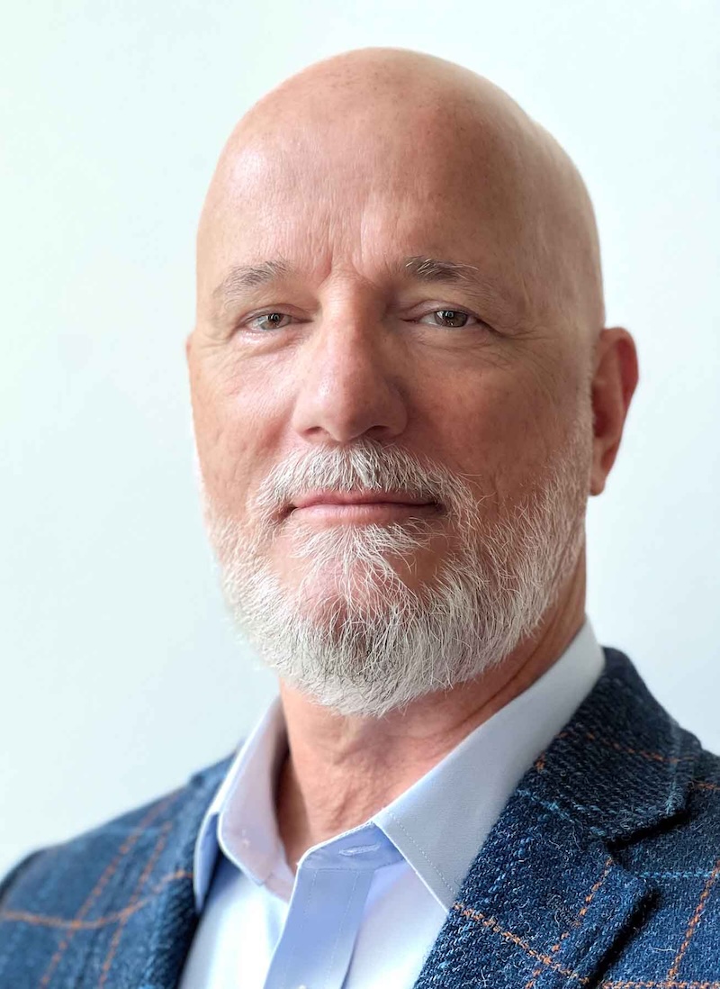 A headshot of Professor Robert Brigham, a person with a shaved head, a thin gray beard, and a blue jacket.