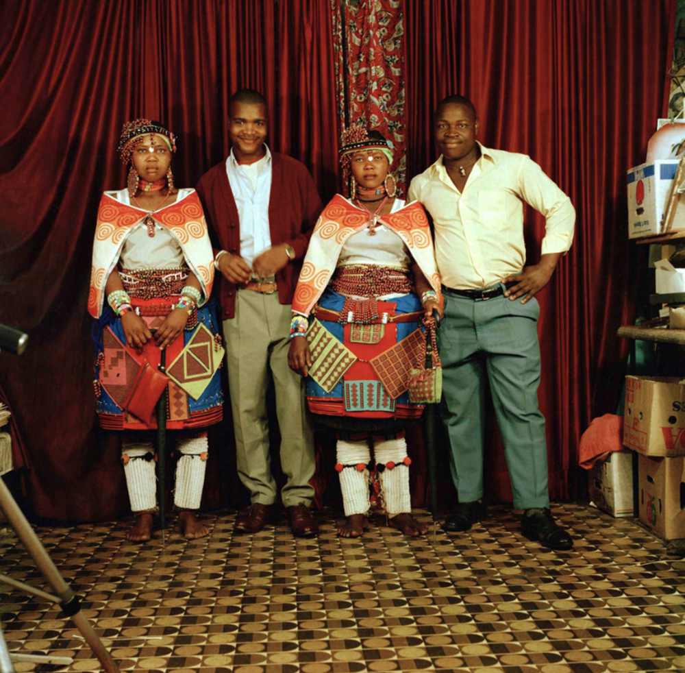 Four people stand against a red velvet curtain. Two of the people are wearing brightly colored clothes.