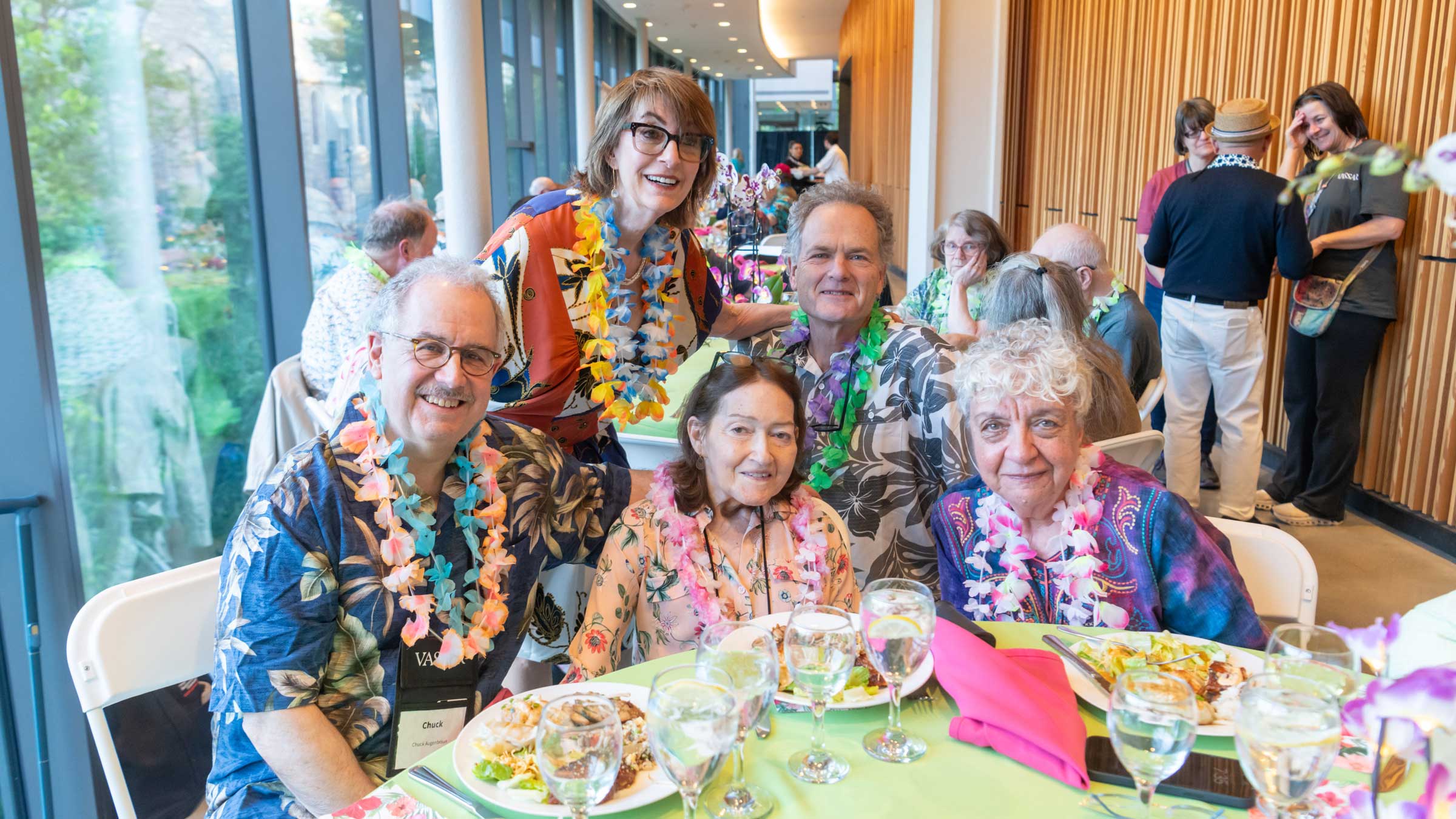 People standing and seated around a table with food wearing tropical print shirts and leis.