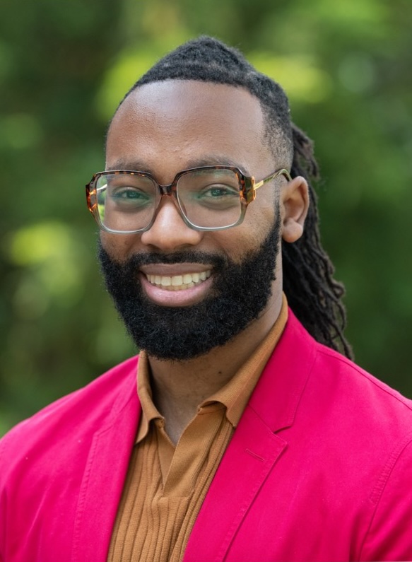 A headshot of Wesley Dixon, a person with long black braided hair, a black beard, glasses, and a bright pink jacket.