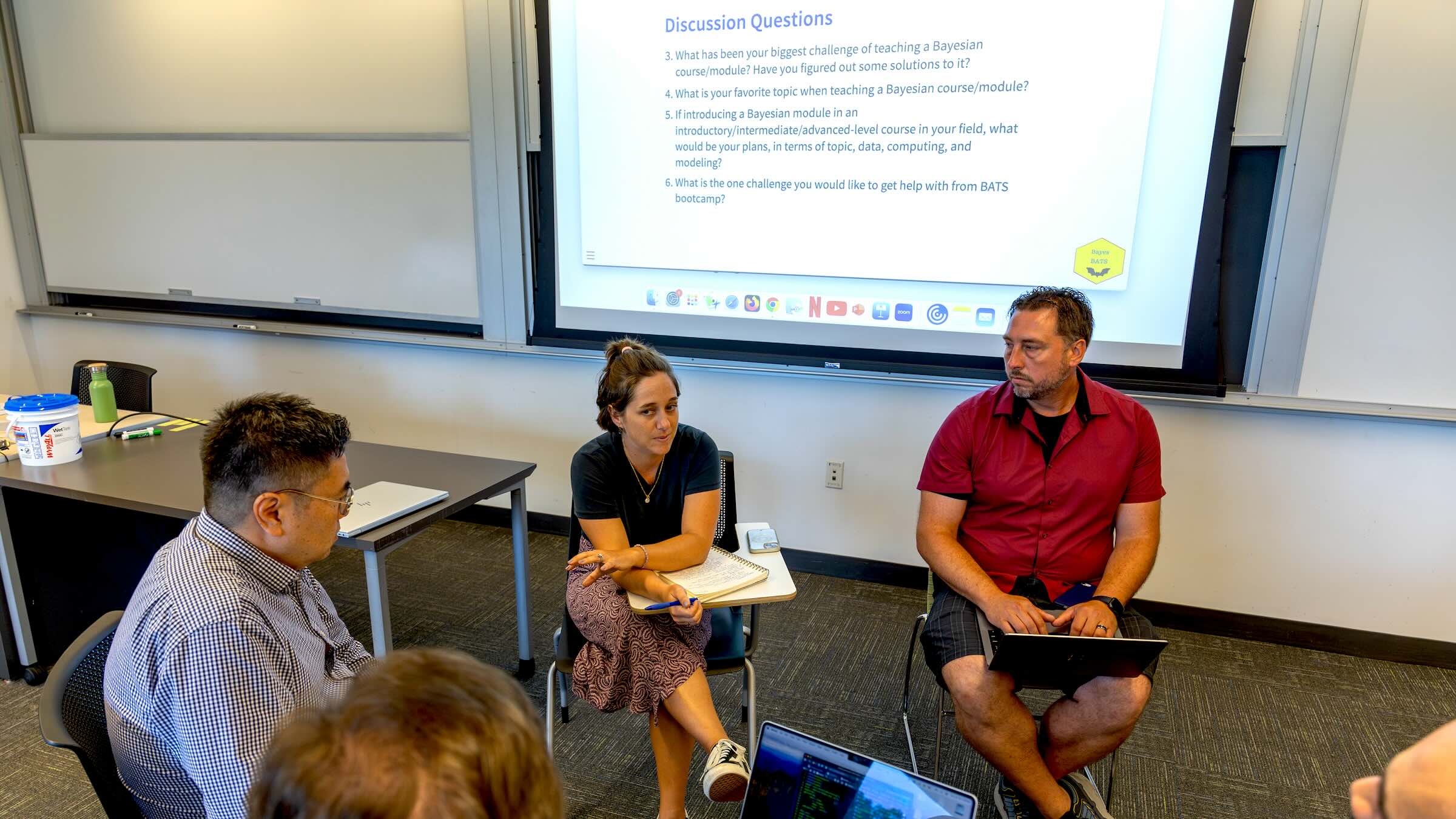 People seated in a circle, some with laptops, in front of a presentation screen, having a discussion.