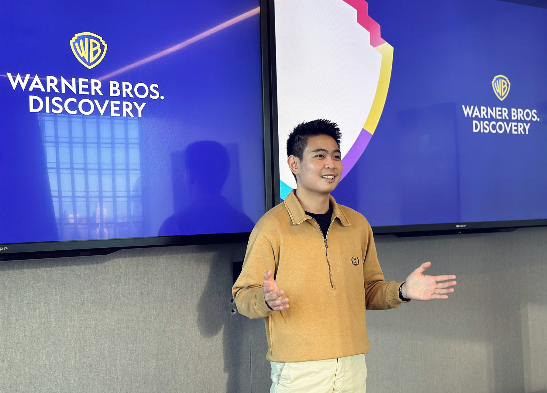 Person standing at the front of a room speaking with display screens in the background that reads, "Warner Brothers Discovery".