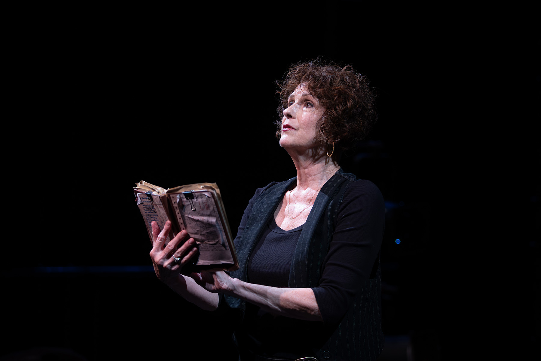 Person standing on a dark stage holding an old book and reading out loud.