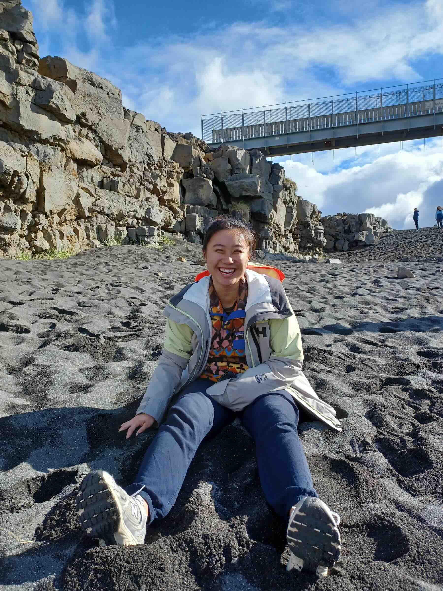 A smiling person sitting in the sand on a beach with a rock cliff and a bridge in the background.