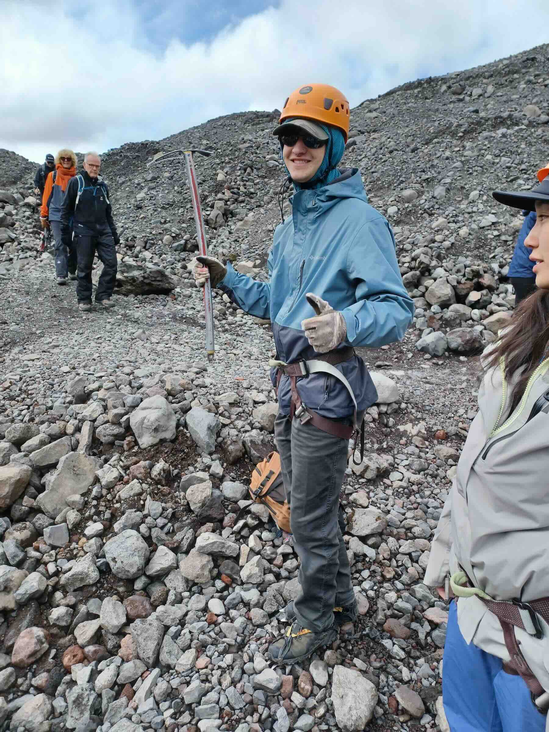 A smiling person in climbing gear holding a pick axe giving a thumbs up.