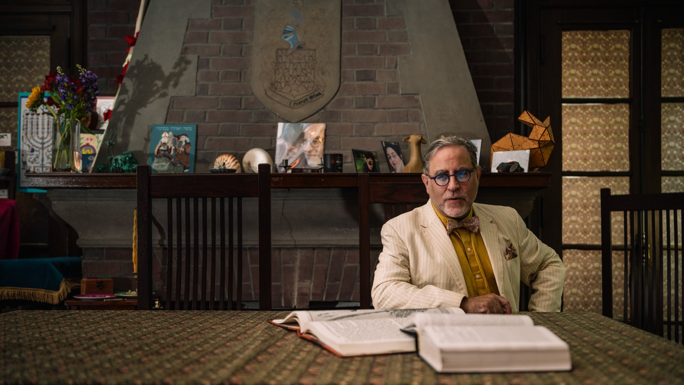 Person in a white suit seated behind a table behind two open books.