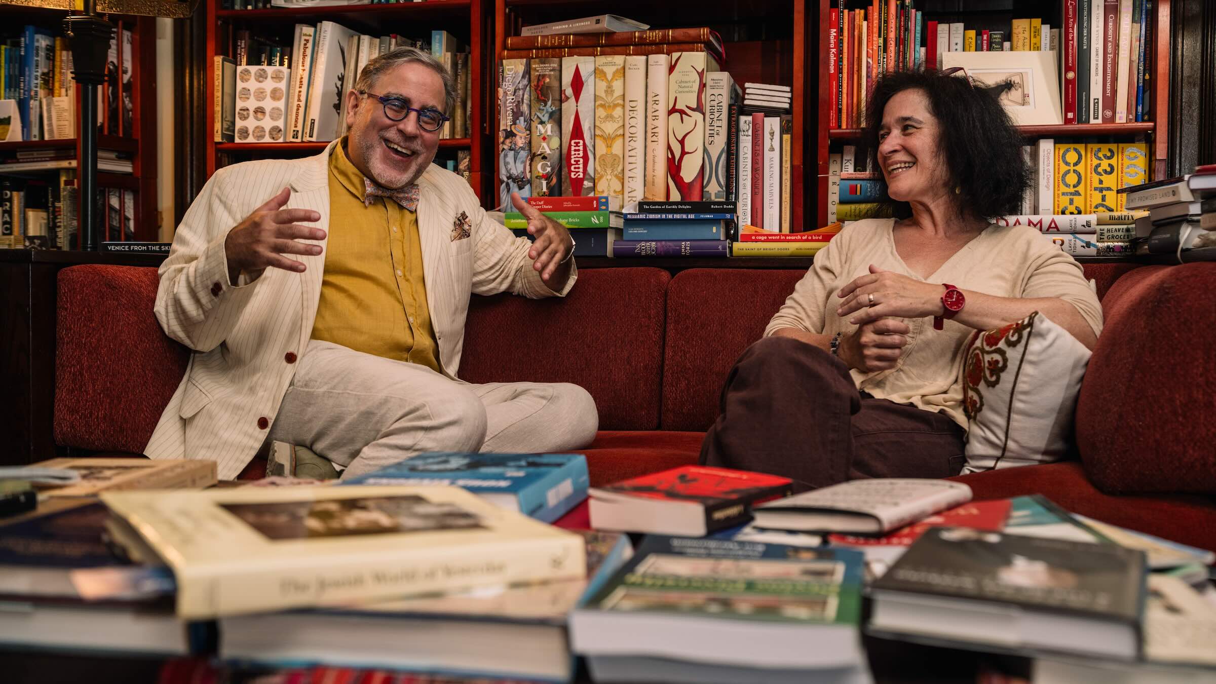 Two people seated on a sofa laughing and smiling while surrounded by books in a bookshelf behind then and a coffee table in front.