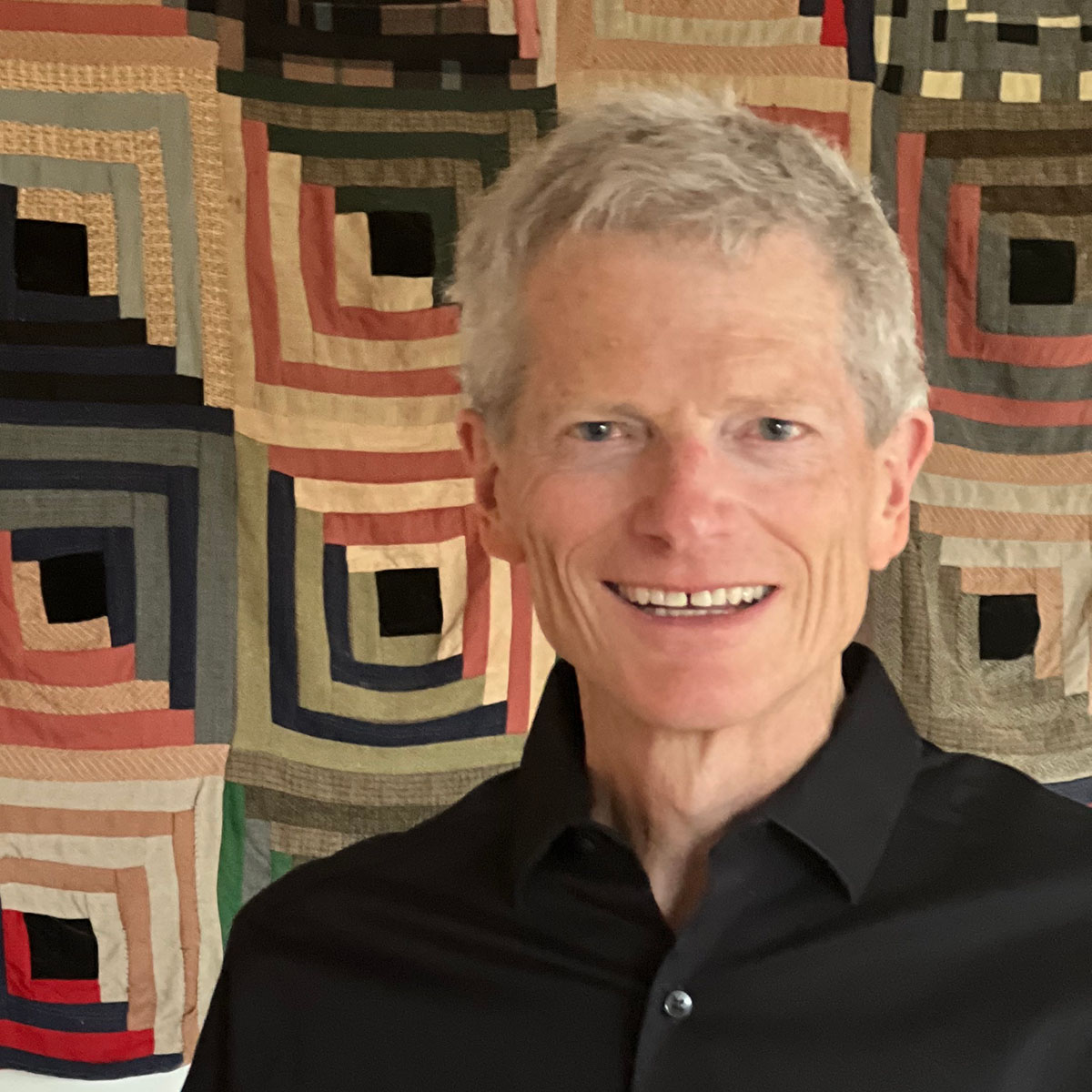 Person with white hair and a black shirt in front of a quilted cover hanging on a wall.