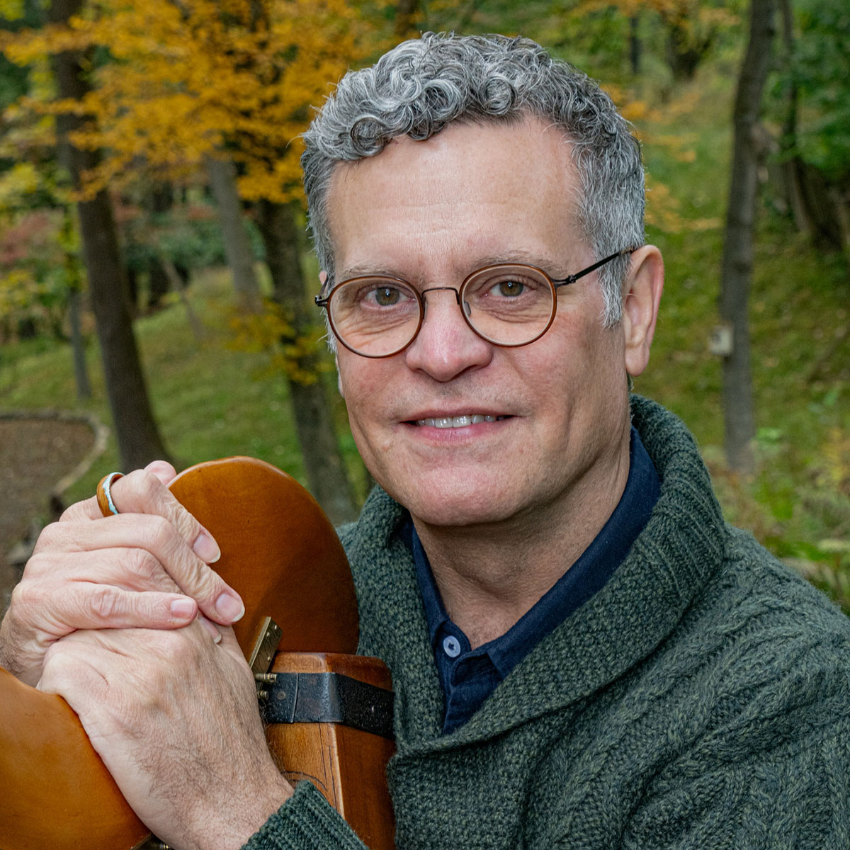 Person with glasses and grey curly hair sitting outside with a forest in the background  holding a harp in their lap.