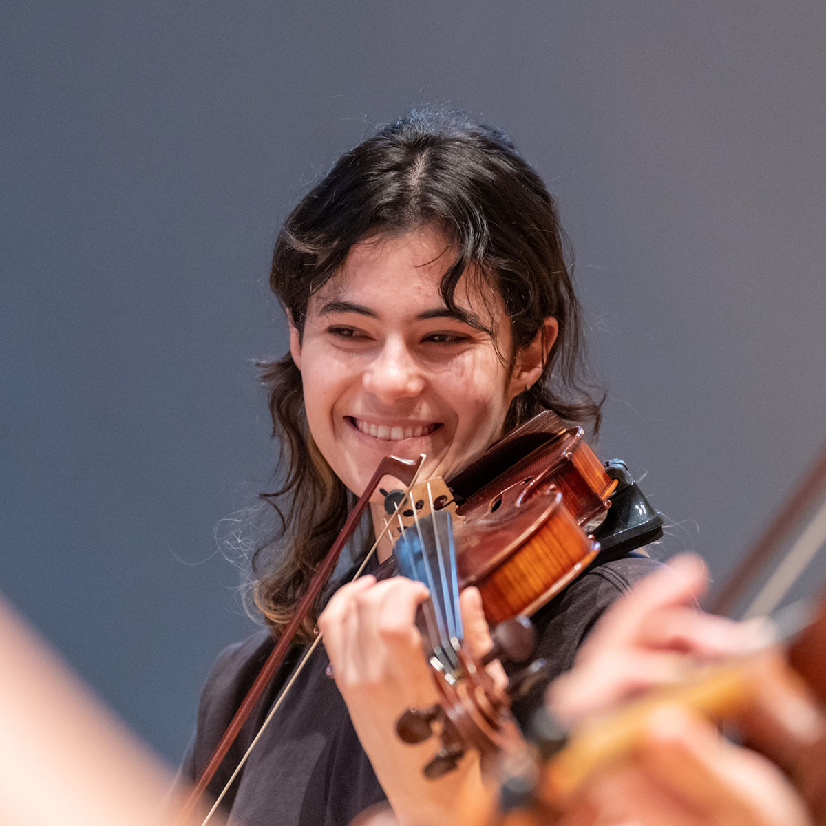 Person with long brown hair on a stage smiling and playing a violin.