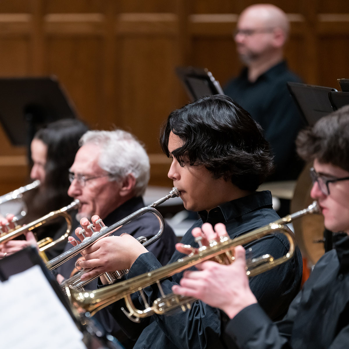 People sitting in a line on stage wearing black and all playing trumpets.