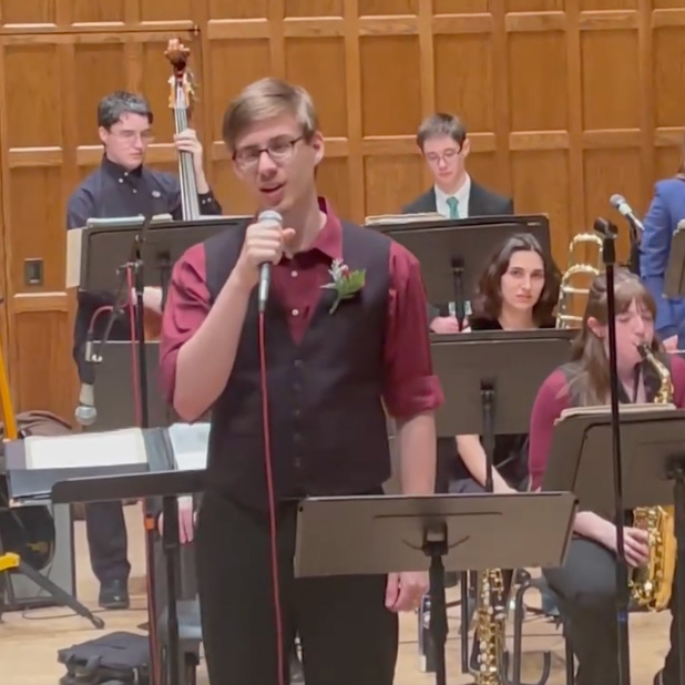 People in an orchestra on a stage with a person in the center sining into a microphone with musicians playing their instruments behind them.