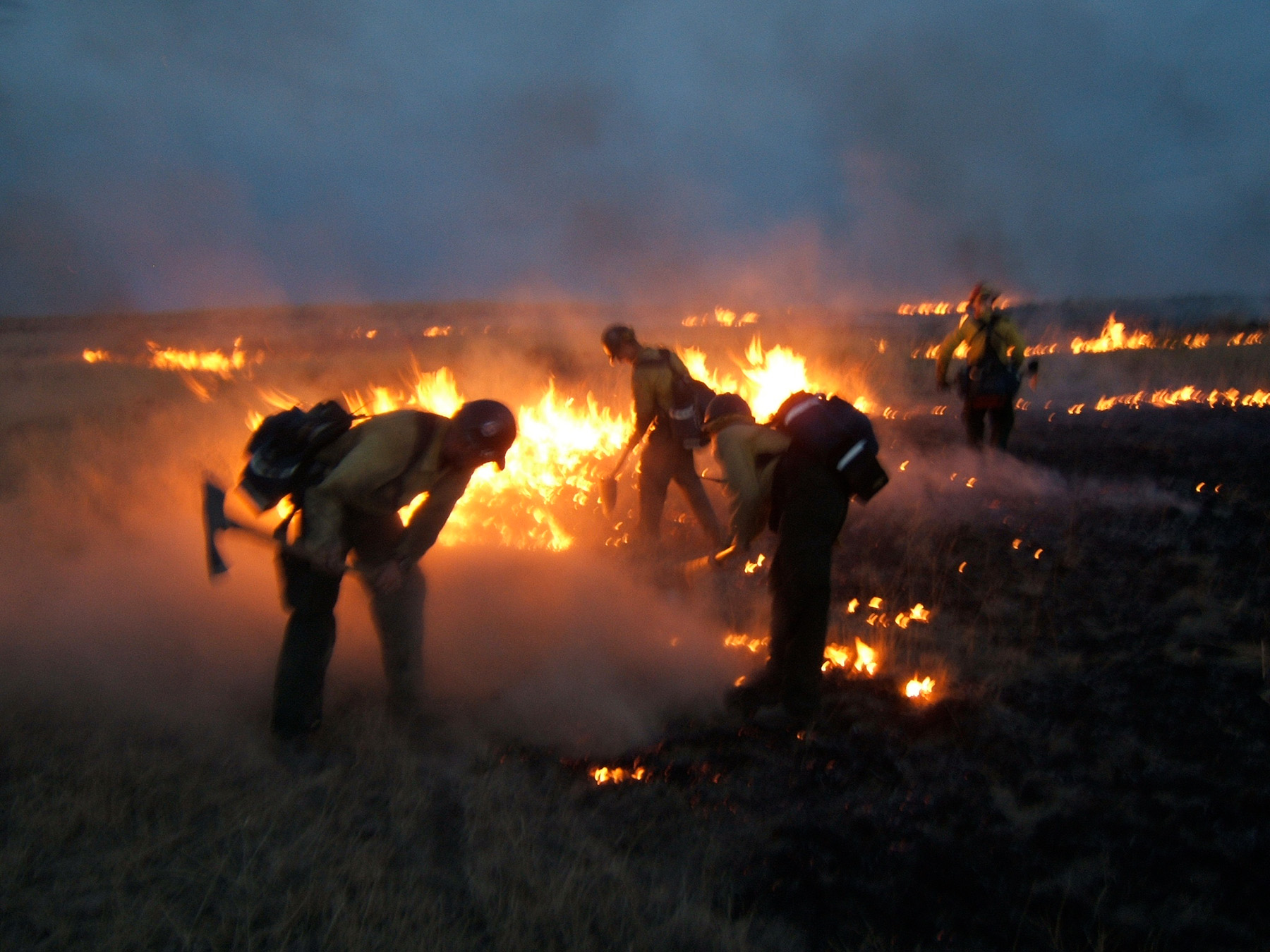 Firefighters fighting an outdoor wildfire with the land on fire in the background with a dark, hazy, smoke-filled sky.