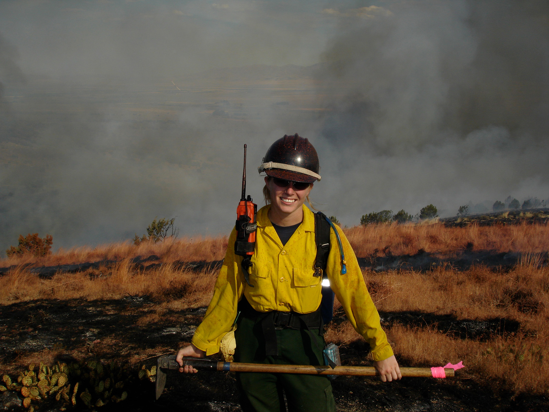 Pictured: Samantha Orient ’05. A person standing with fire-fighting gear (caution-yellow shirt and overalls) holding hatchet/shovel.