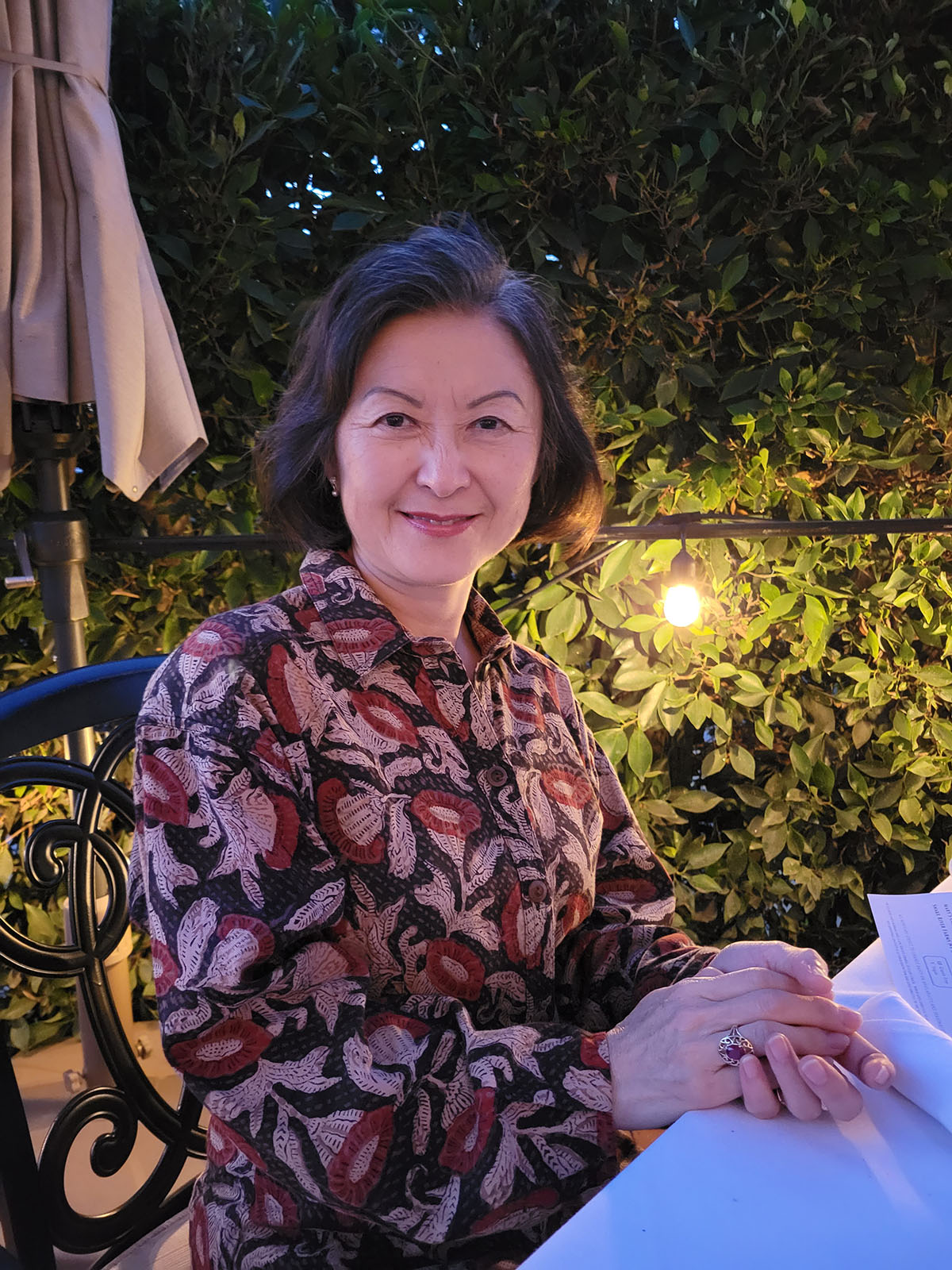 Person with black hair and a flower dress sitting at an outdoor table with trees in the background.