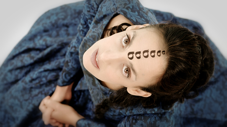 Person looking towards the sky in a large blue dress with letters drawn on her forehead.