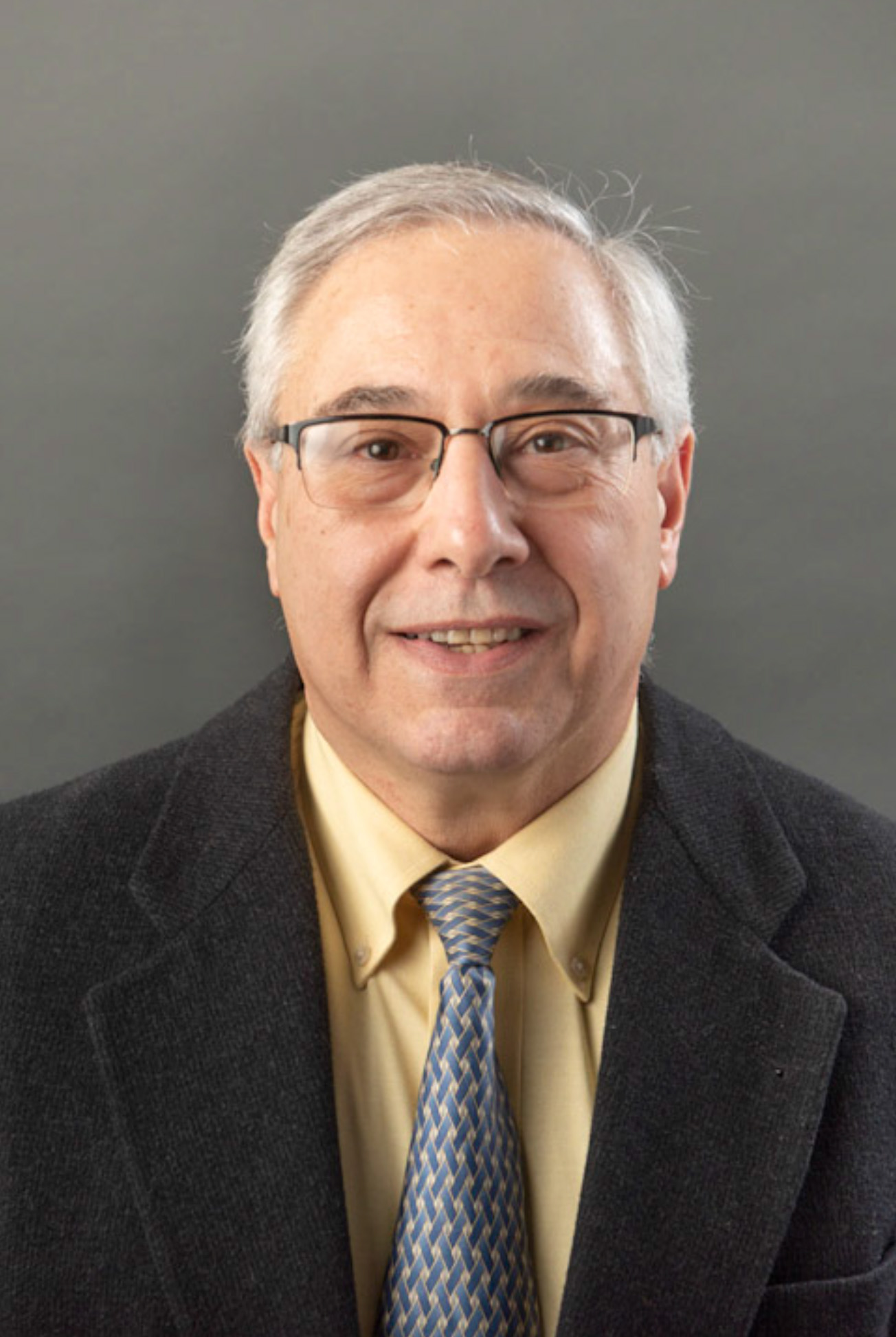 Portrait of Anthony Scaduto. Person with grey hair and glasses wearing a black suit and tie with a yellow shirt.