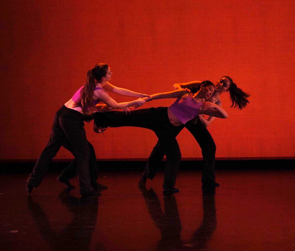 Three students dance on a dark stage, the room lit a dim red.