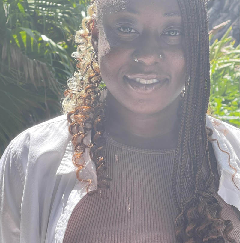 Fanta Wague, a student with a white shirt and long dark hair, sits smiling in front of greenery.