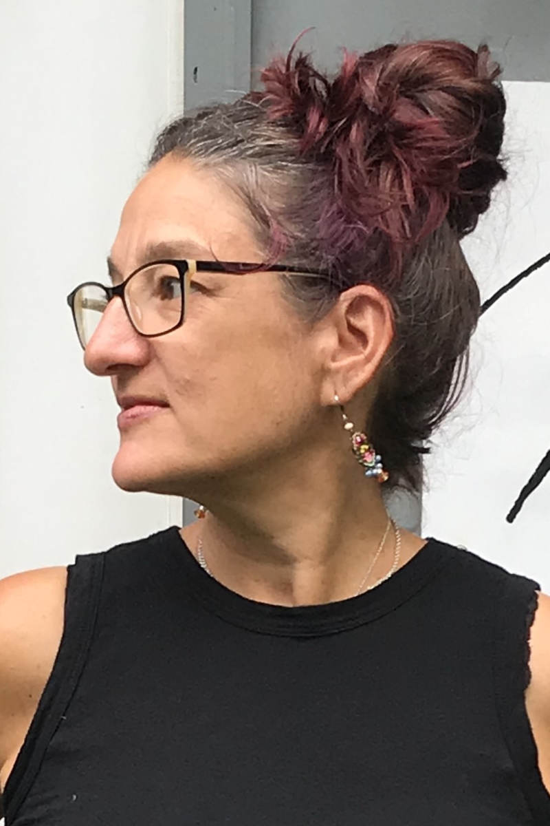 A headshot of Jessie Levey, a person with long dark hair in a bun, glasses, and a black tank top. They are seen in profile, looking to their right.