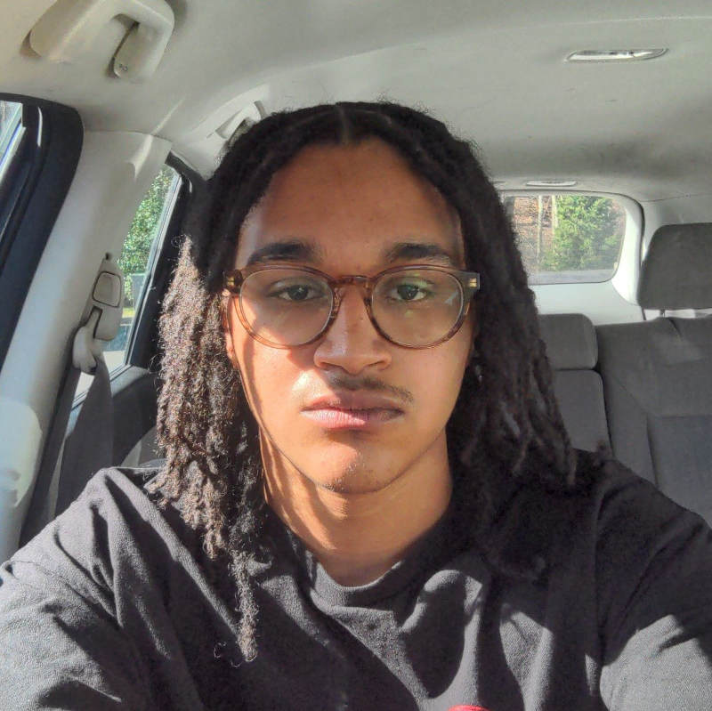 A portrait of Kwabena Adae, a student with long dark curly hair, glasses, and a thin mustache, sitting in the front seat of a car.