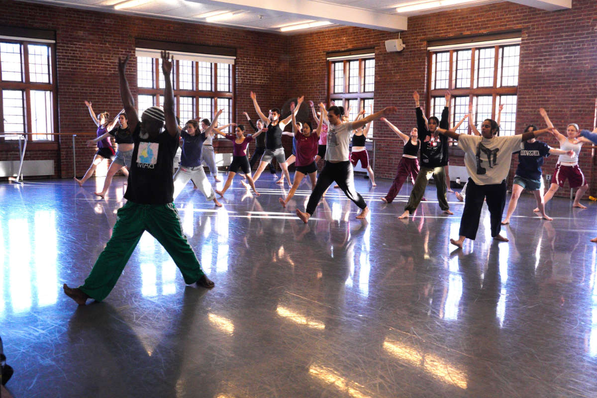 A person teaching a dance class in a large brick studio filled with other people.