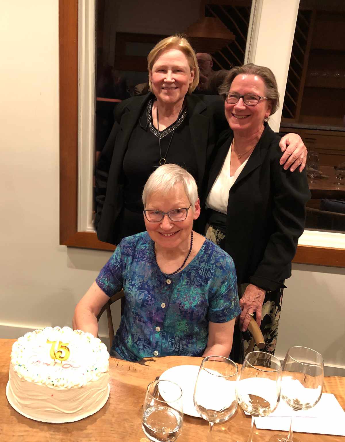 Three people in a room, smiling at the camera. One of the people is seated at a table, on which is placed a cake.