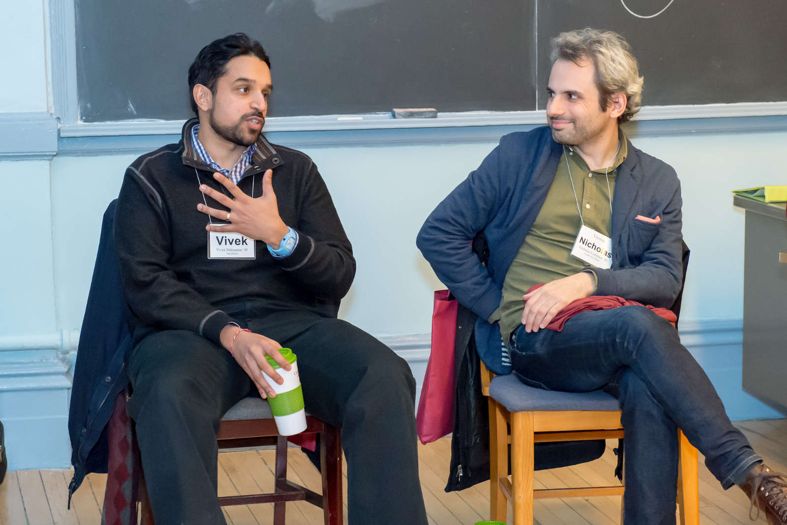 Two people seated on chairs in front of a blackboard, talking.