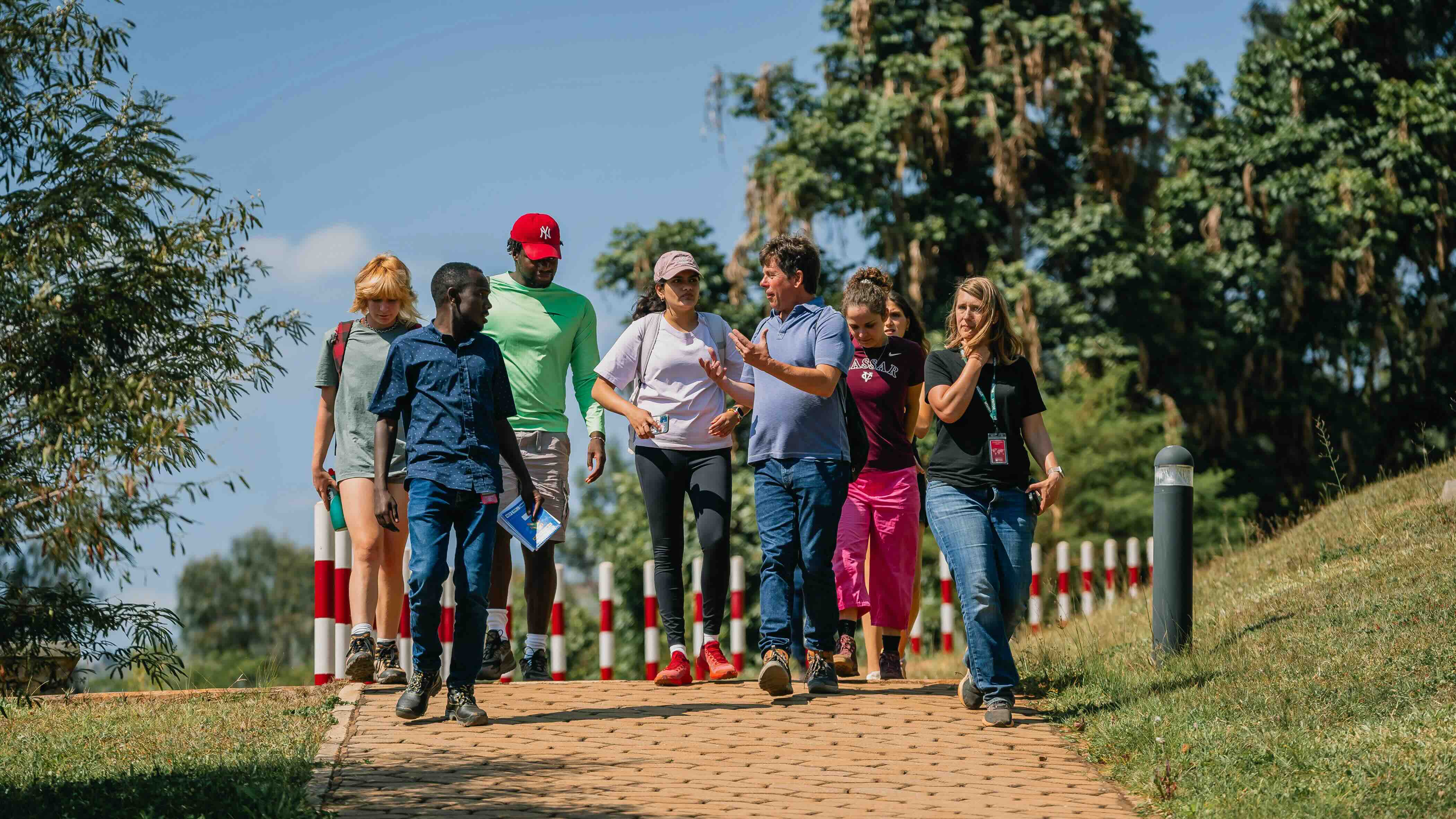 A group of people walking and talking on a paved path through a grassy area in bright midday sunlight.