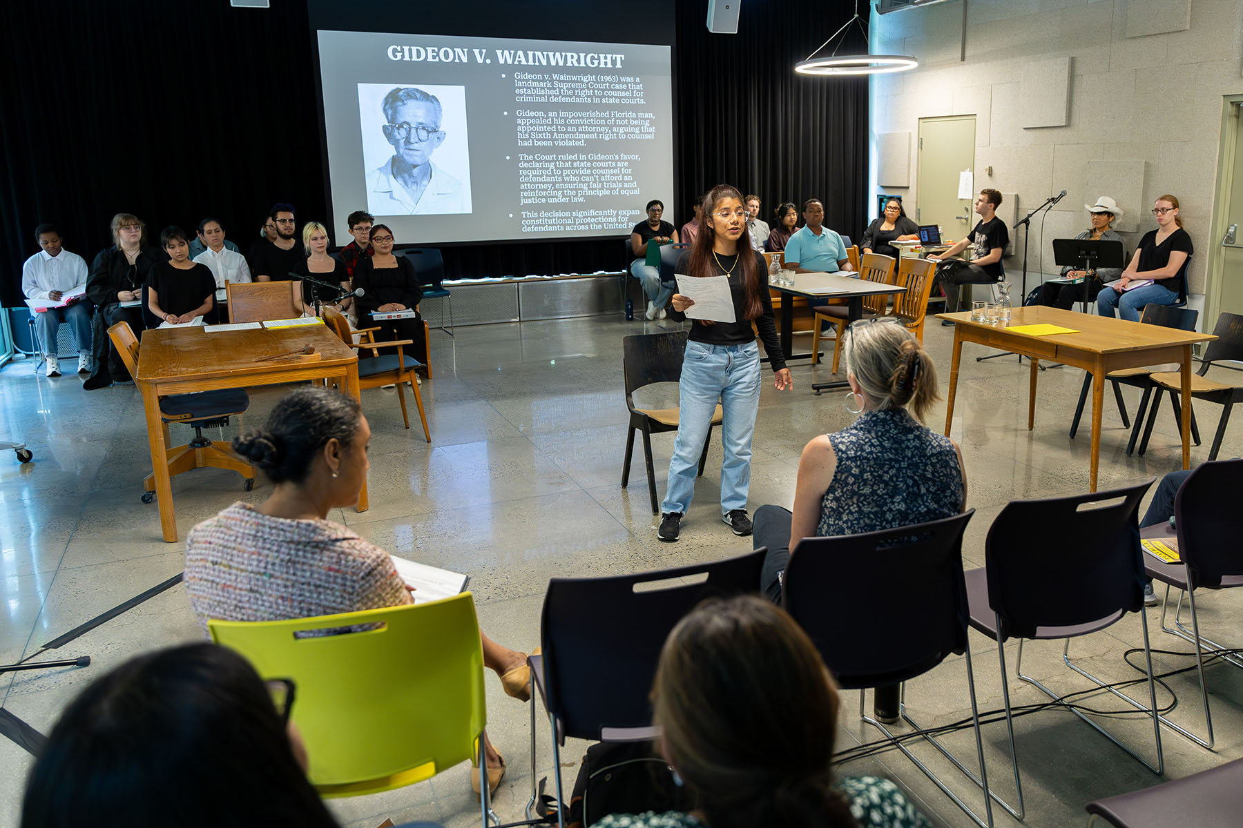 A speaker standing in the center of a room holding a piece of paper with people seated at desks in a square around the speaker. There is a projection screen in the background.