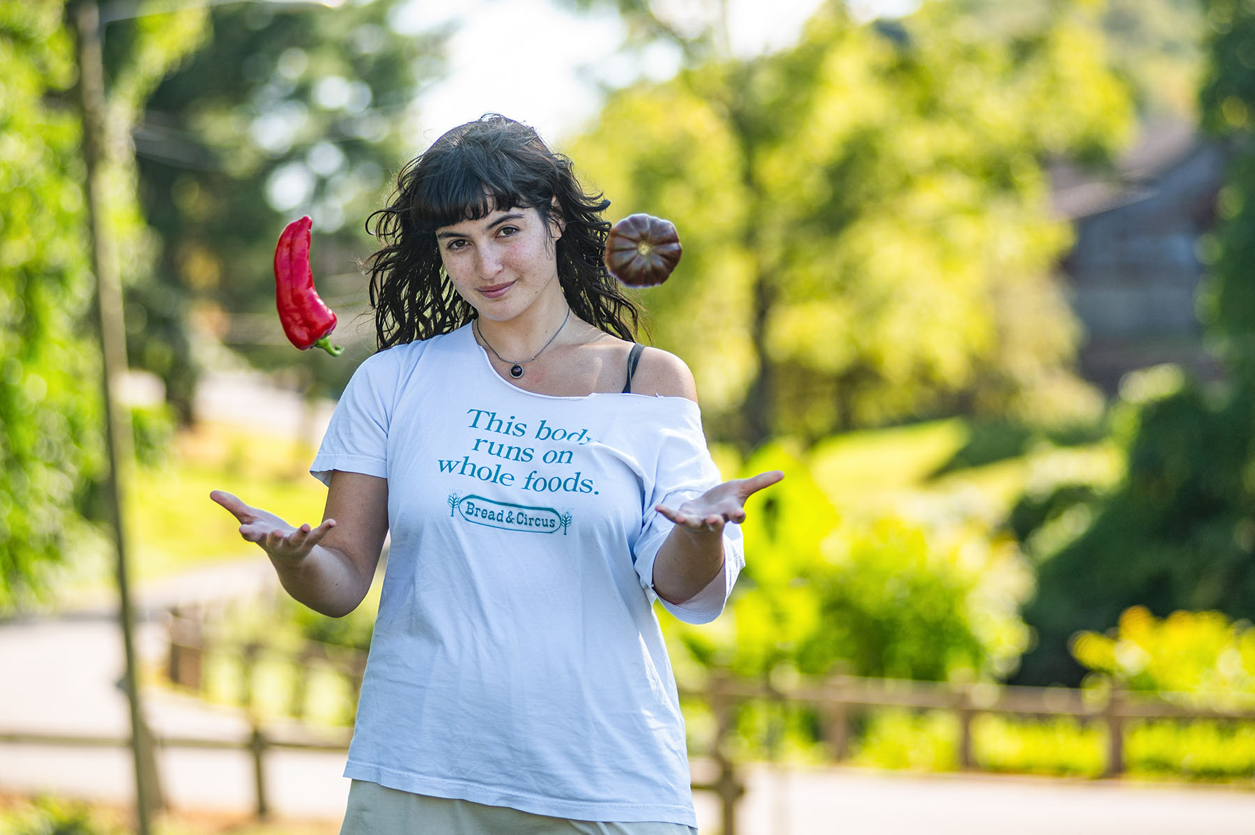 Esther Cull-Kahn wearing a white printed t-shirt and throwing a couple of red peppers in the air with trees in the background.