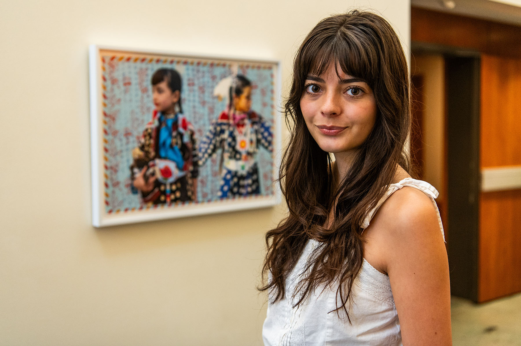 Julia Pippenger with long dark hair and wearing a white strappy shirt, standing next to Indigenous artwork of two young people. 