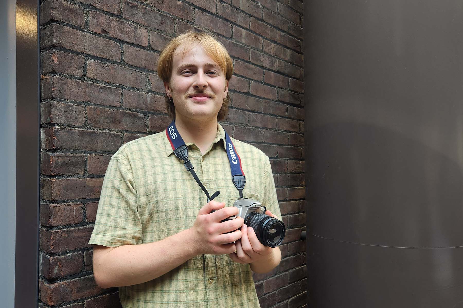 Ben Richardson wearing a green plaid shirt and holding a camera strapped around his neck while standing in front of a brick wall.