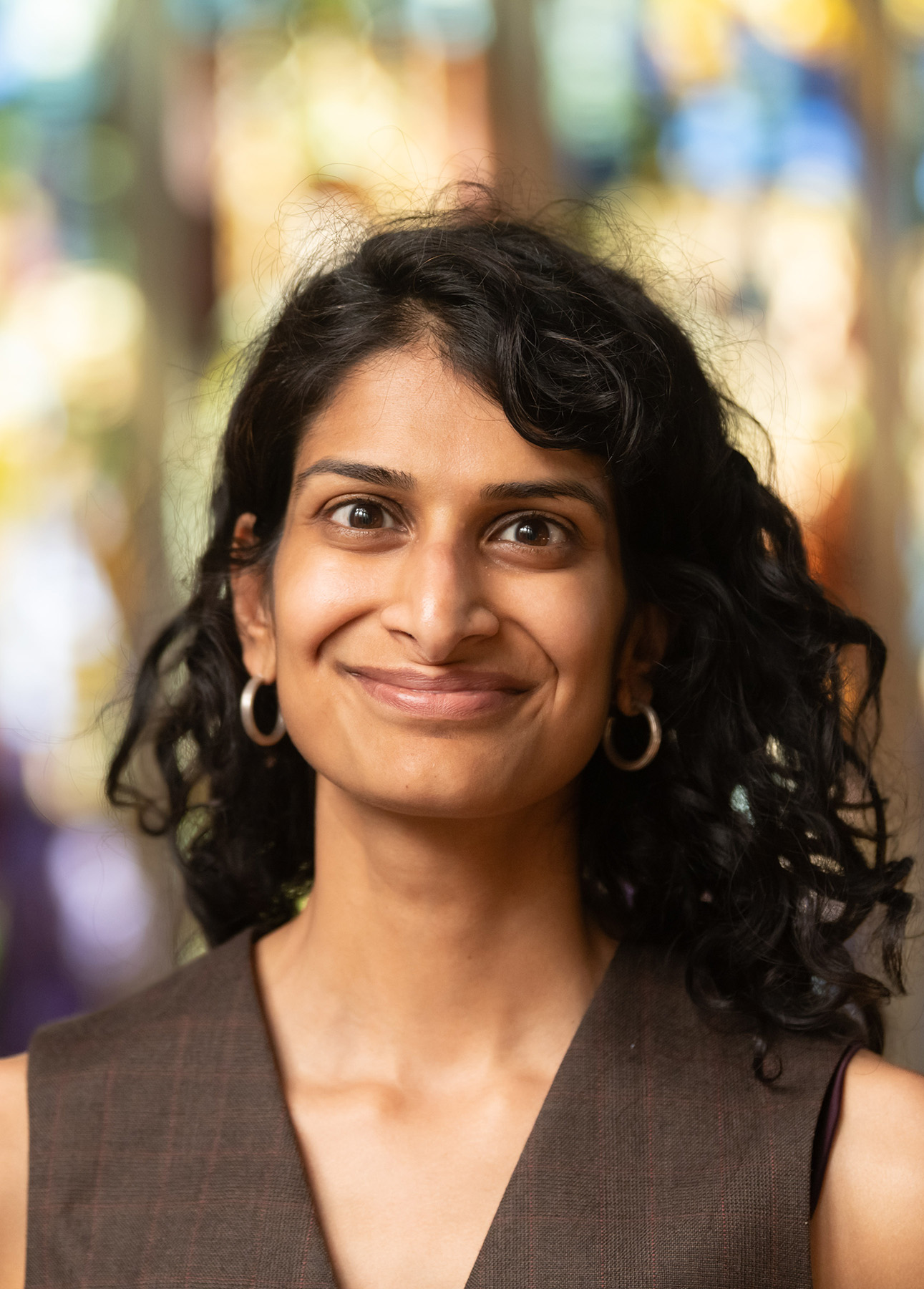 Portrait of a person with long black hair and olive brown blouse smiling with a blurry stained glass window in the background.