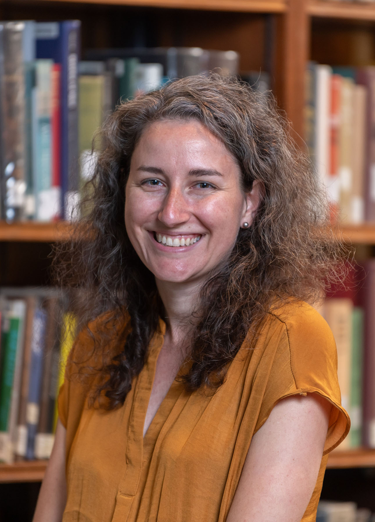 Person with long curly brown hair standing in front of a book case.